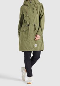 Parka verte à capuche avec fermeture éclair à l'avant, cordons de serrage, multiples poches, portée sur un pantalon noir et des baskets blanches.