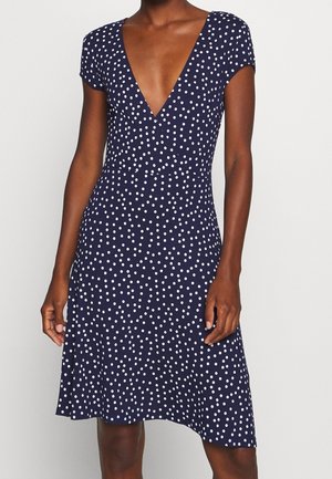 Kobieta ubrana w granatową sukienkę z krótkimi rękawami w białe kropki z głębokim dekoltem w kształcie litery V na jednolitym tle.