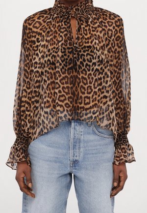 Blouse transparente imprimé léopard avec un col haut froncé, lien devant en forme de trou de serrure, poignets smockés, portée avec un jean bleu clair.