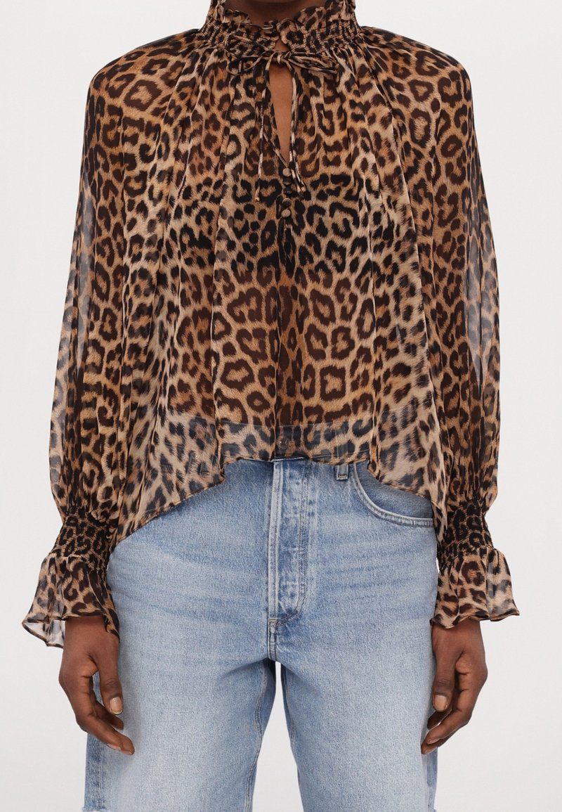 Blouse transparente imprimé léopard avec un col haut froncé, lien devant en forme de trou de serrure, poignets smockés, portée avec un jean bleu clair.