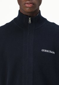 Mørkeblå hettejakke med glidelås og ribbestrikket krage, laget av strikket stoff. Har en hvit "GUESS JEANS" logo på venstre bryst.