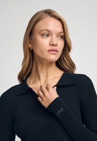 Svart ribbad tröja med V-halsringning, med långa ärmar och broderad detalj på ärmslutet. Slät yta och figurnära design.