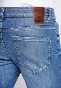 Jeans i mellanblå denim med texturerad yta. Innehåller en brun läderlapp och en bakficka med synlig sömnad.