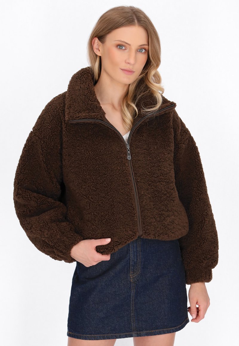 Brun fleecejacka med hög krage, hel dragkedja och ballongsärmar. Bärs över en denimkjol och har en mjuk, texturerad yta.