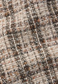 Gros plan sur un tissu texturé avec un motif tissé en fils beige, marron, noir et blanc créant un motif à carreaux.