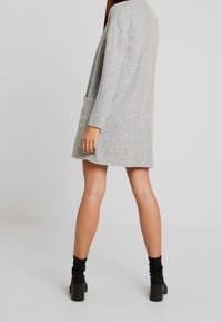Cárdigan gris de punto con corte suelto, mangas largas y un bolsillo delantero. Textura suave y diseño hasta la rodilla. Combinado con botas negras hasta el tobillo.