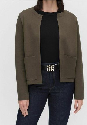 Kvinde iført en olivengrøn åben jakke med store lommer over en sort top, mørke jeans og et sort bælte med en gylden dekorativ spænde.