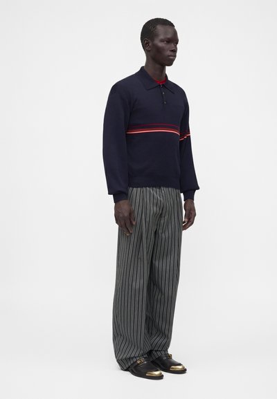 Homme portant un polo bleu marine à manches longues avec des rayures rouges, un pantalon ample rayé noir et blanc, et des chaussures noires avec des bouts dorés, debout devant un fond blanc.