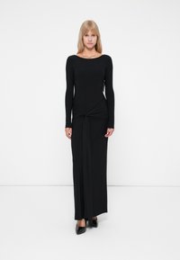 Musta pitkähiha maxi-mekko, jossa on istuva bodice, solmittu yksityiskohta vyötäröllä ja laskostettu hame. Sileä, joustava kangas.