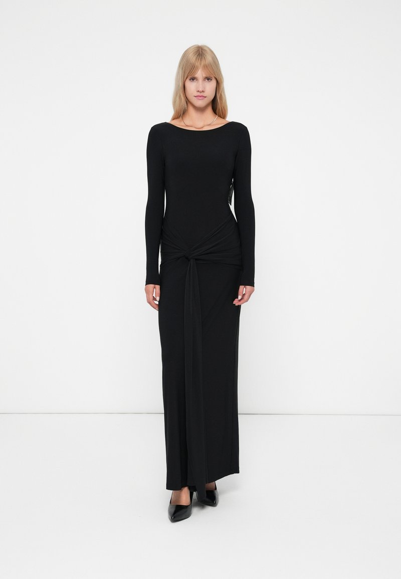 Musta pitkähiha maxi-mekko, jossa on istuva bodice, solmittu yksityiskohta vyötäröllä ja laskostettu hame. Sileä, joustava kangas.