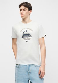 Νέος άνδρας που φοράει λευκό μπλουζάκι με γραφικό παλαιού τύπου ραδιοφώνου και μικροφώνου και το κείμενο "Η Φωνή των Θαραλλέα, Alpha Radio" και ανοιχτόχρωμο τζιν.