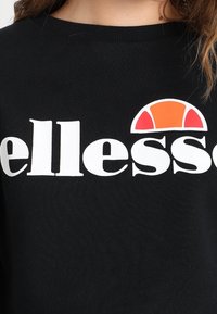 Černá mikina s velkým bílým logem "ellesse" a oranžovým a žlutým polokruhem navrchu. Materiál vypadá měkký a texturovaný.