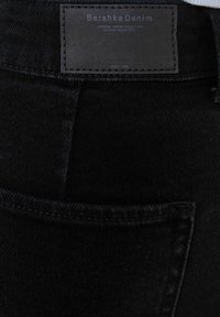Mustakangas, jossa on lähikuva takataskusta. Suorakulmainen etiketti, jossa lukee "Bershka Denim" kohokuvioituna ja ompelutiedot näkyvissä.