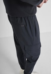 Persona che indossa pantaloni cargo blu scuro con vita e orlo elasticizzati, con tasche laterali e posteriori, abbinati a sneaker bianche.