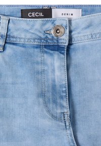 Jean en denim bleu clair avec fermeture par bouton argenté et étiquette visible portant la mention "Cecil Denim" à l'intérieur de la ceinture.