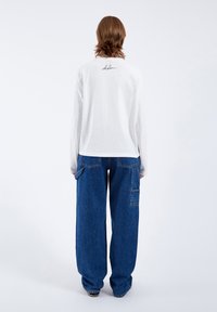 White long-sleeve shirt with a rounded hem and black embroidered logo on the back, paired with loose-fitting blue denim pants featuring pockets.