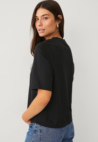 Schwarzes T-Shirt mit kurzen Ärmeln und rundem Ausschnitt. Aus glattem Stoff gefertigt, mit lockerer Passform und leicht abgerundetem Saum. Kombiniert mit blauen Jeans.