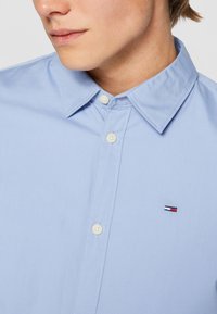 Camicia azzurra chiaro con colletto classico, bottoni bianchi e un piccolo logo ricamato sul lato sinistro del petto. Tessuto dalla texture liscia.