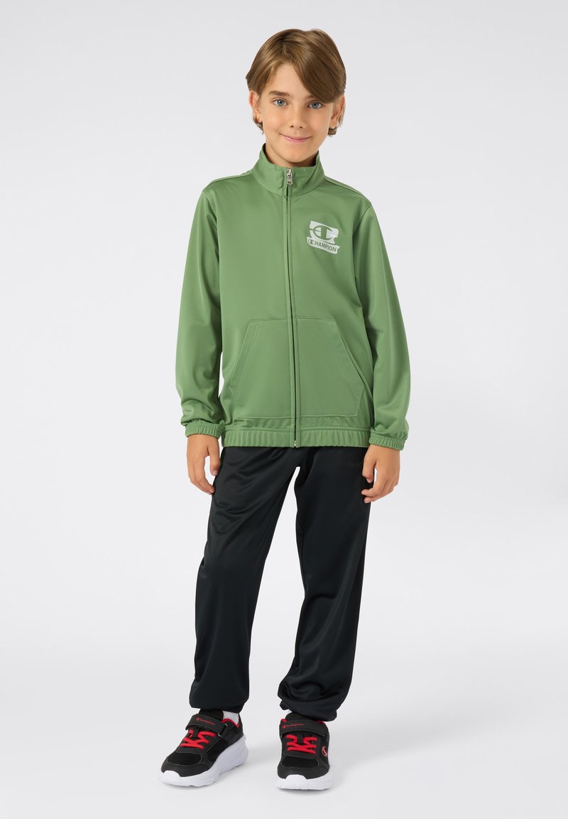 Groene sportjack met hoge kraag, rits en voorzakken. Gecombineerd met zwarte broek en zwarte sneakers met rode accenten.