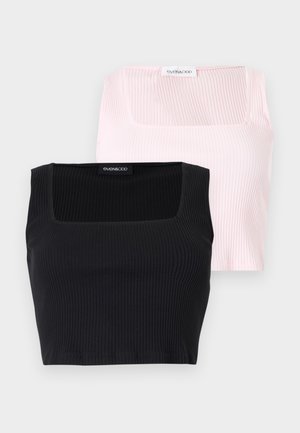 Ung kvinde med bølget hår iført en lyserød ærmeløs crop top, sorte cykelshorts med høj talje og sorte flipflops, stående mod en hvid baggrund.