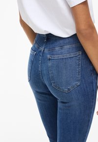 Pantalones vaqueros de denim azul ajustados con cintura alta y dos bolsillos traseros, que presentan una textura suave y detalles de costura sutiles.