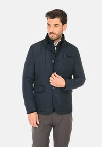 Giacca trapuntata blu navy con collo alla coreana, chiusura con zip e bottoni, tasche frontali e una tasca sul petto. Realizzata con un materiale liscio e leggero.