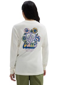 Person trägt ein cremefarbenes Langarmshirt mit buntem Blumendesign, Friedenstaube und dem Schriftzug "Vans Peaceful Bliss Since 1966" auf dem Rücken.