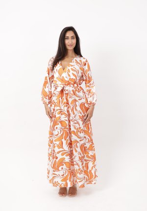Vrouw met lang donker haar, die een witte maxijurk draagt met oranje bloemmotieven en een taillekoord, blootsvoets staand op een witte achtergrond.