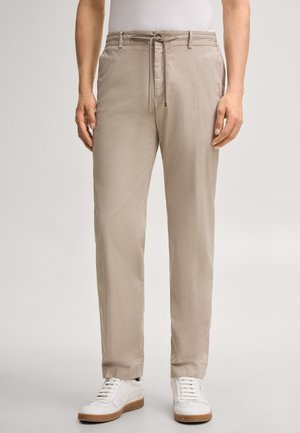 Beige broek met trekkoord, gedragen met witte sneakers en een wit topje, weergegeven van taille tot voeten tegen een effen achtergrond.