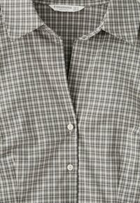 Chemise à carreaux grise et blanche boutonnée avec col, présentant deux boutons blancs visibles et une étiquette de la marque Stradivarius à l'intérieur du col.