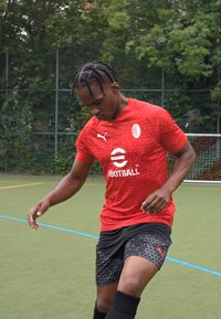 Maglia atletica rossa con un motivo tessutato, pantaloni corti neri con un design geometrico e calze nere. Logo Puma e testo "FOOTBALL" visibili.