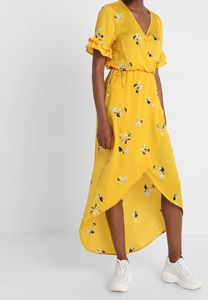 Gele bloemen wikkeljurk met geruite korte mouwen en een hoog-laag zoom, gedragen met witte chunky sneakers.