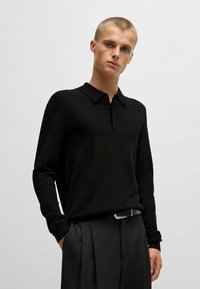 Schwarzes Langarm-Polohemd mit Kragen und einem halben Reißverschluss vorne. Das Material hat eine glatte Textur. Kombiniert mit maßgeschneiderten schwarzen Hosen.