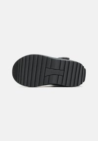 Suela de zapatilla de goma negra con textura estriada y un logo prominente en el centro. Cuenta con un fondo plano para ofrecer tracción y estabilidad.