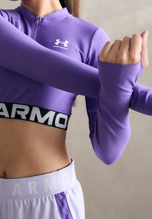 Frau streckt die Arme, trägt ein lila Under Armour cropped Langarmtop und weiße Shorts mit lila Streifen vor einfarbigem Hintergrund.