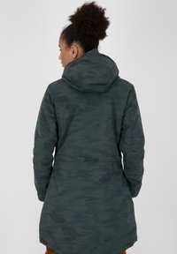 Giacca camouflage verde con cappuccio, caratterizzata da una vita con cordino, maniche lunghe e tessuto texturizzato. Include una toppa sulla spalla.