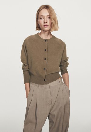 Brown knitted cardigan with buttons, featuring a rounded neckline and ribbed cuffs, paired with light beige pleated trousers.
