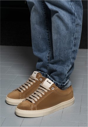 Person wearing light brown suede sneakers with beige soles and laces, paired with rolled-up blue denim jeans on a tiled floor.