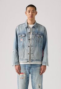 Veste en denim bleu clair à coupe classique, avec deux poches poitrine, des détails usés et une étiquette rouge sur le côté.