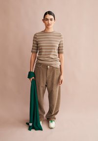 Bruine gestreepte t-shirt gecombineerd met losse beige corduroy broek. Model houdt een groene sjaal vast, draagt witte sneakers met groene accenten.