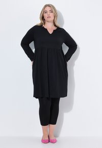 Robe noire à manches longues avec un décolleté en V, taille froncée, associée à des leggings noirs et des ballerines roses. Texture de tissu lisse.
