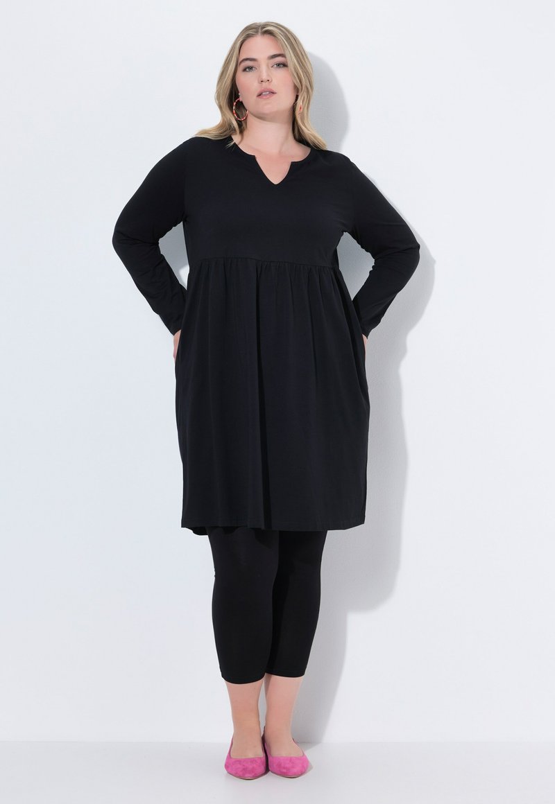 Robe noire à manches longues avec un décolleté en V, taille froncée, associée à des leggings noirs et des ballerines roses. Texture de tissu lisse.