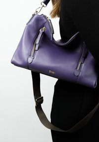 Sac à main en cuir violet avec des zips dorés et des accents en pampille. Il comprend une bandoulière marron amovible et une finition lisse et texturée.