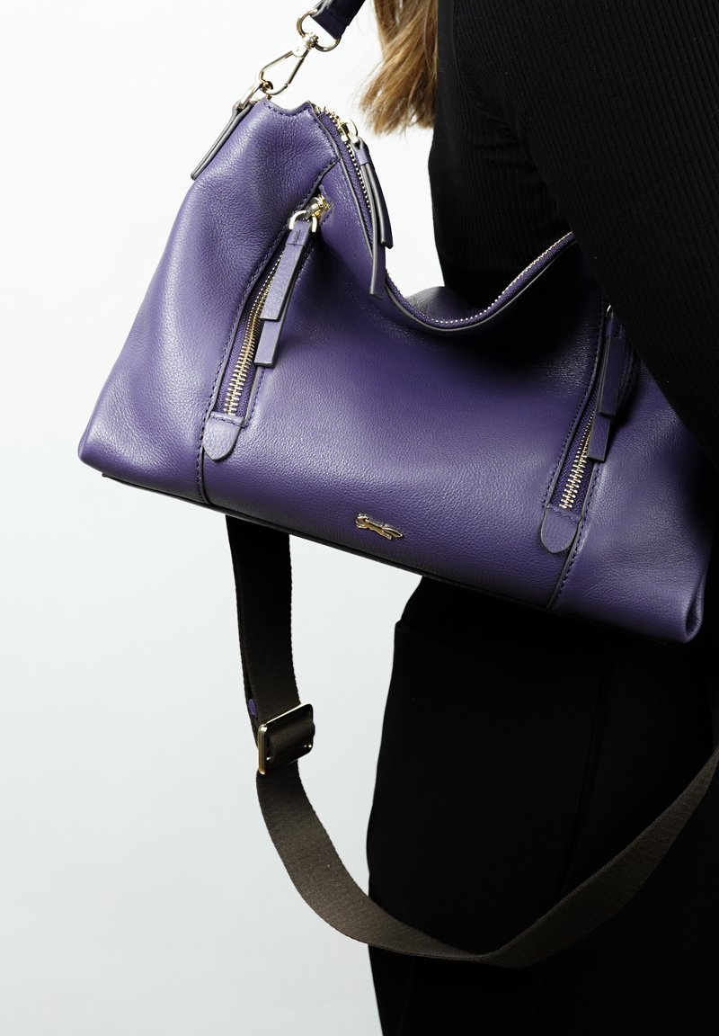 Sac à main en cuir violet avec des zips dorés et des accents en pampille. Il comprend une bandoulière marron amovible et une finition lisse et texturée.