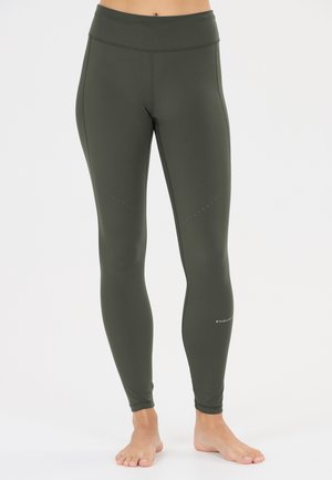 Vrouw draagt olijfgroene, hooggetailleerde sportleggings, staat op blote voeten met benen iets uit elkaar op een effen lichte achtergrond.
