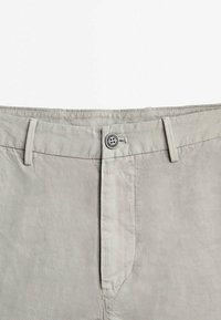 Ceinture de pantalon gris clair avec fermeture par bouton, passants pour ceinture et coutures visibles sur fond uni.