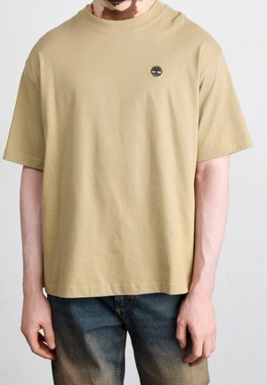 Mand iført beige kortærmet t-shirt med lille sort trælogo på brystet og falmede blå jeans mod ensfarvet baggrund.