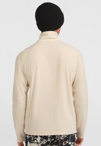 Beige fleecejakke med høj krave og lange ærmer, glat tekstur, parret med en sort strikket hue.