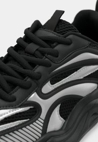 Nahaufnahme eines schwarzen und grauen Sport Sneakers, der strukturiertes Material, dicke Schnürsenkel und Details der skulptierten Gummisohle zeigt.