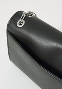 Sac en simili cuir noir avec une texture lisse, doté d'un accent en chaîne en métal et d'une forme structurée. Détail de poche latérale visible.